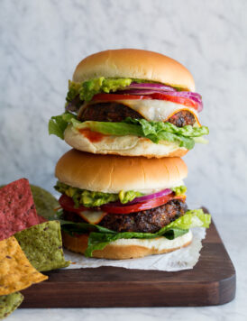 Two black bean burgers in buns with lettuce, tomatoes, smashed avocado, red onion, and cheese.