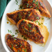 Row of blackened chicken breast on a white oval platter over a marble surface with a green cloth to the side.