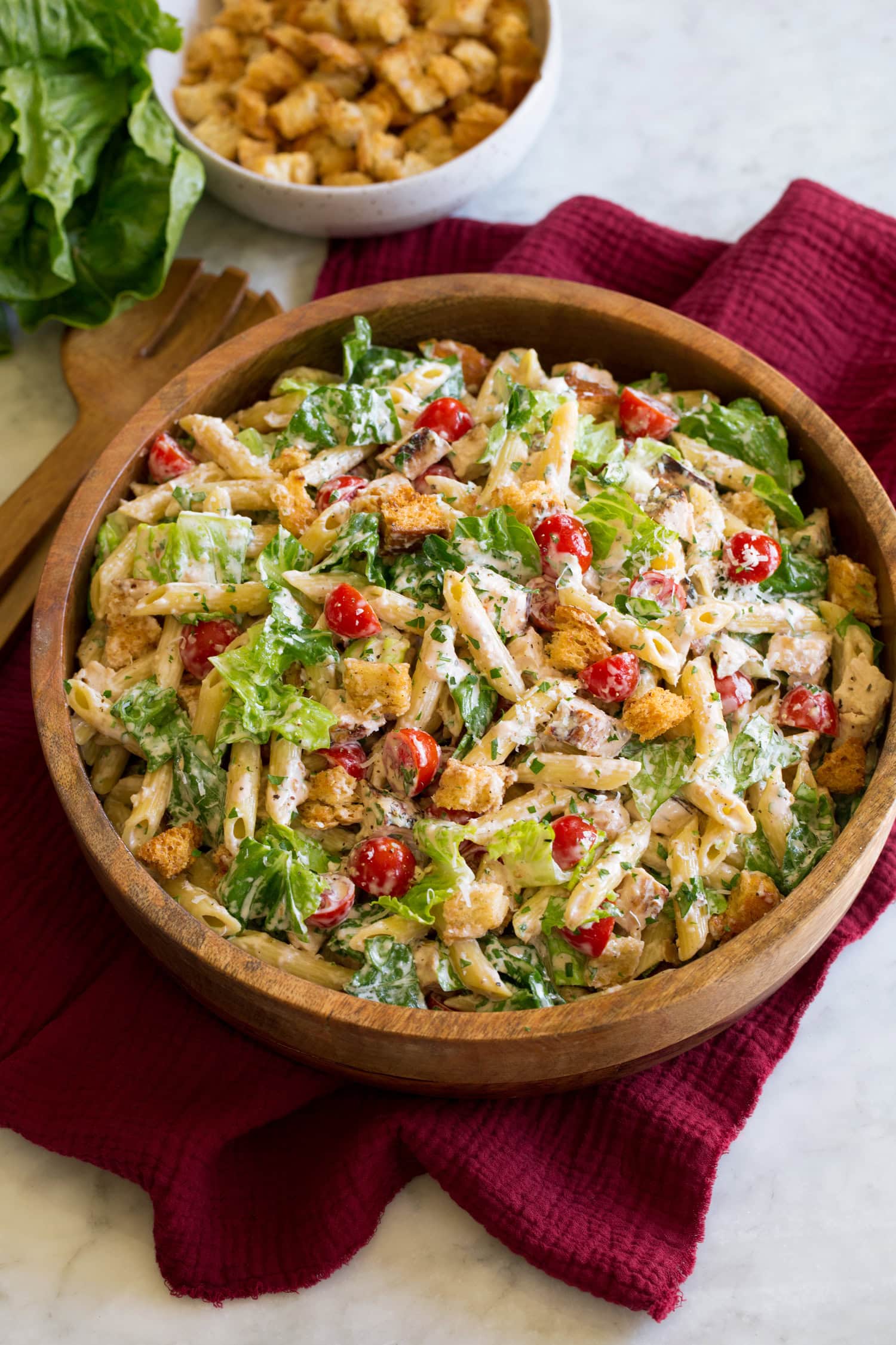 Chicken caesar pasta salad shown in a brown wooden serving bowl over a red cloth on a white marble surface.