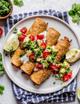 Taquitos on a white plate topped with sour cream, tomatoes, avocados and cilantro. Lime wedges are set of to the side and plate is set on a blue striped napkin.