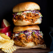 Photo: Two BBQ Pulled Chicken Sandwiches stacked on a wood platter. Apples slices and potato chips are shown to the side as serving suggestions.