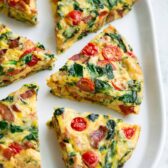 Six frittata wedges shown on a white serving platter set over a marble surface. Frittata is filled with bacon, spinach, tomatoes and swiss.