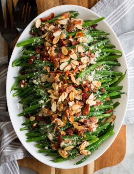 Green Bean Almondine. Sauteed green beans are shown lined up in a row on a platter with toasted almonds, crumbled fried bacon and grated parmesan are sprinkled over the top.