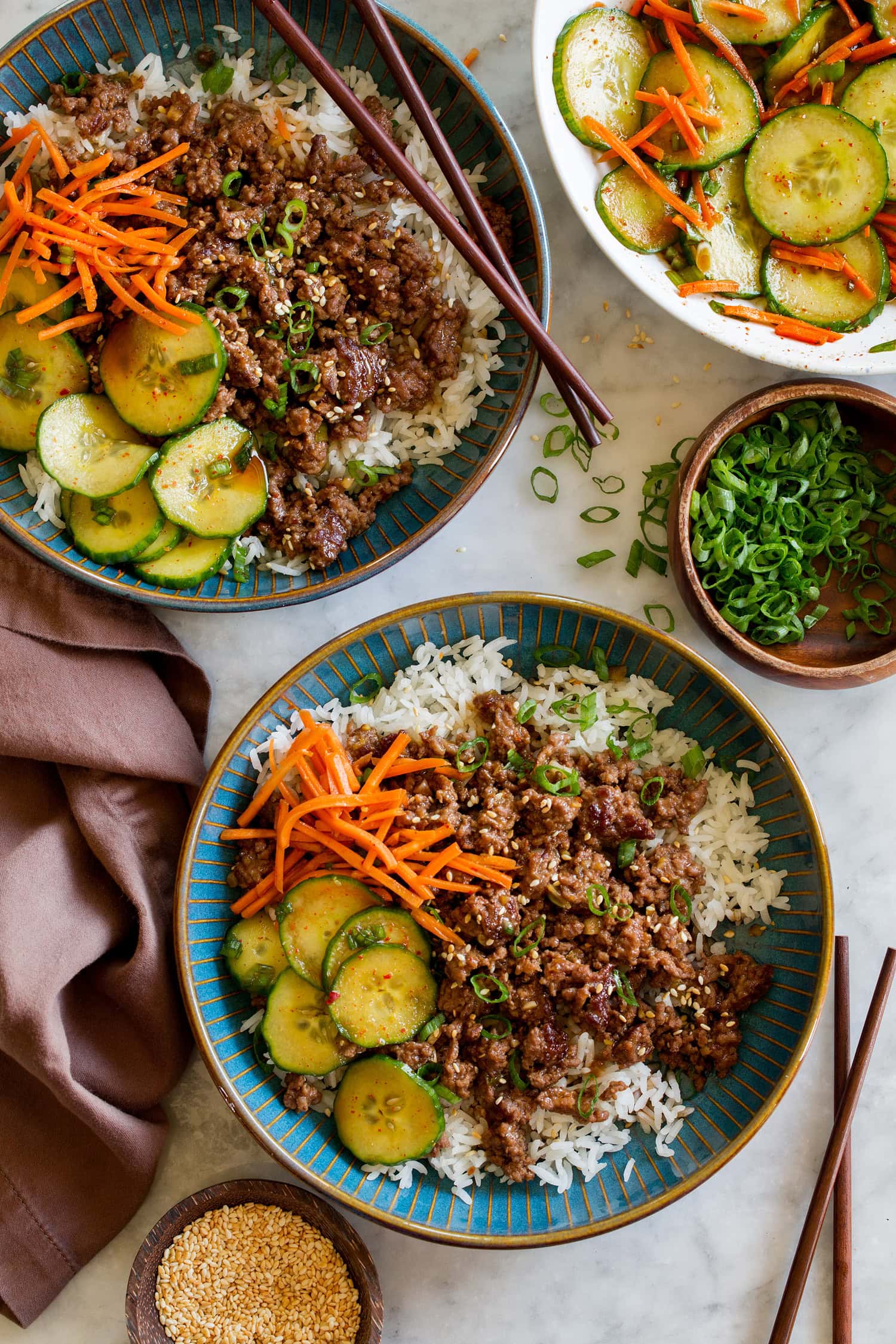 Korean beef bowls shown with serving suggestion of sides of Korean vegetables including cucumbers and carrots.