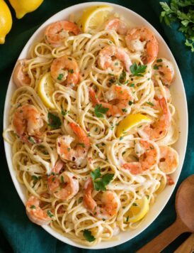 Shrimp pasta with creamy lemon sauce on a white oval serving platter shown from above. Platter is resting on an aqua colored cloth and lemons are shown to the side.