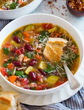 Image of serving of minestrone soup in a white bowl set over white plate. Soup is garnished with parmesan and topped with bread.