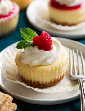 Mini cheesecake shown on a dessert plate unwrapped from cupcake liner. It is topped with whipped cream, fresh raspberry and mint.