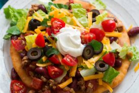 Homemade navajo taco on a plate. Made with homemade fry bread, ground beef and bean filling, lettuce, tomatoes, olives and sour cream.