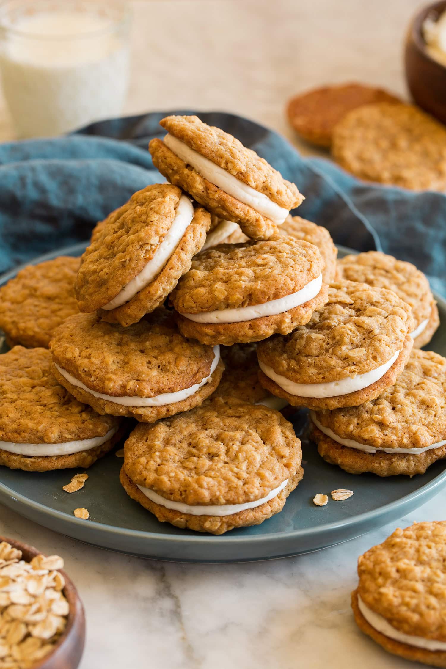 Stack of oatmeal cream pies on a blue plate with a blue cloth do the side and a glass of milk.