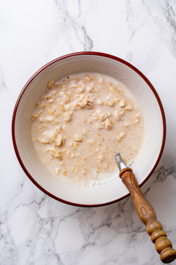 A spoon with a wooden handle rests in a bowl of mixed overnight oats ingredients.