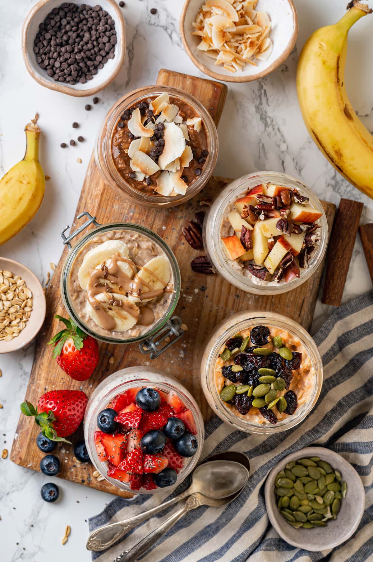 Overhead view of overnight oats in mason jars, five different ways, on a wooden platter surrounded by bowls of toppings and add-ins.