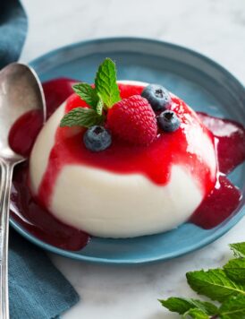 Photo: Single serving of a disk of Panna Cotta shown on a blue plate covered with raspberry sauce, fresh blueberries and mint.