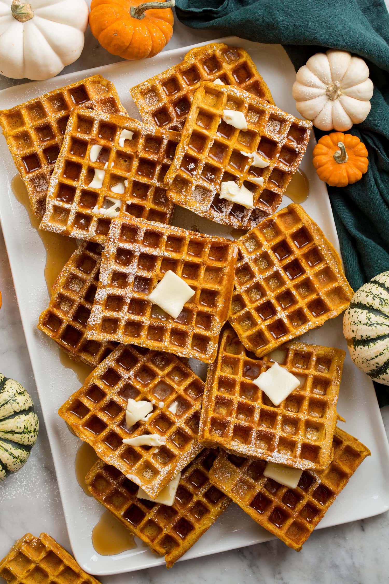 Pumpkin waffles layered on a white rectangular platter covered with syrup and butter, shown from above with pumpkins to the side.