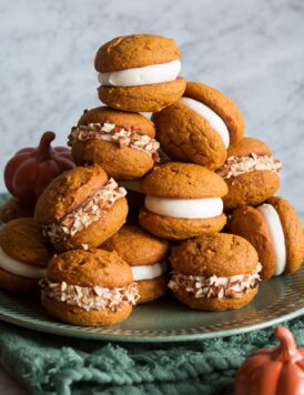 Pumpkin Whoopie Pies stacked on a large plate. They are filled with cream cheese frosting and some are covered with chopped pecans.