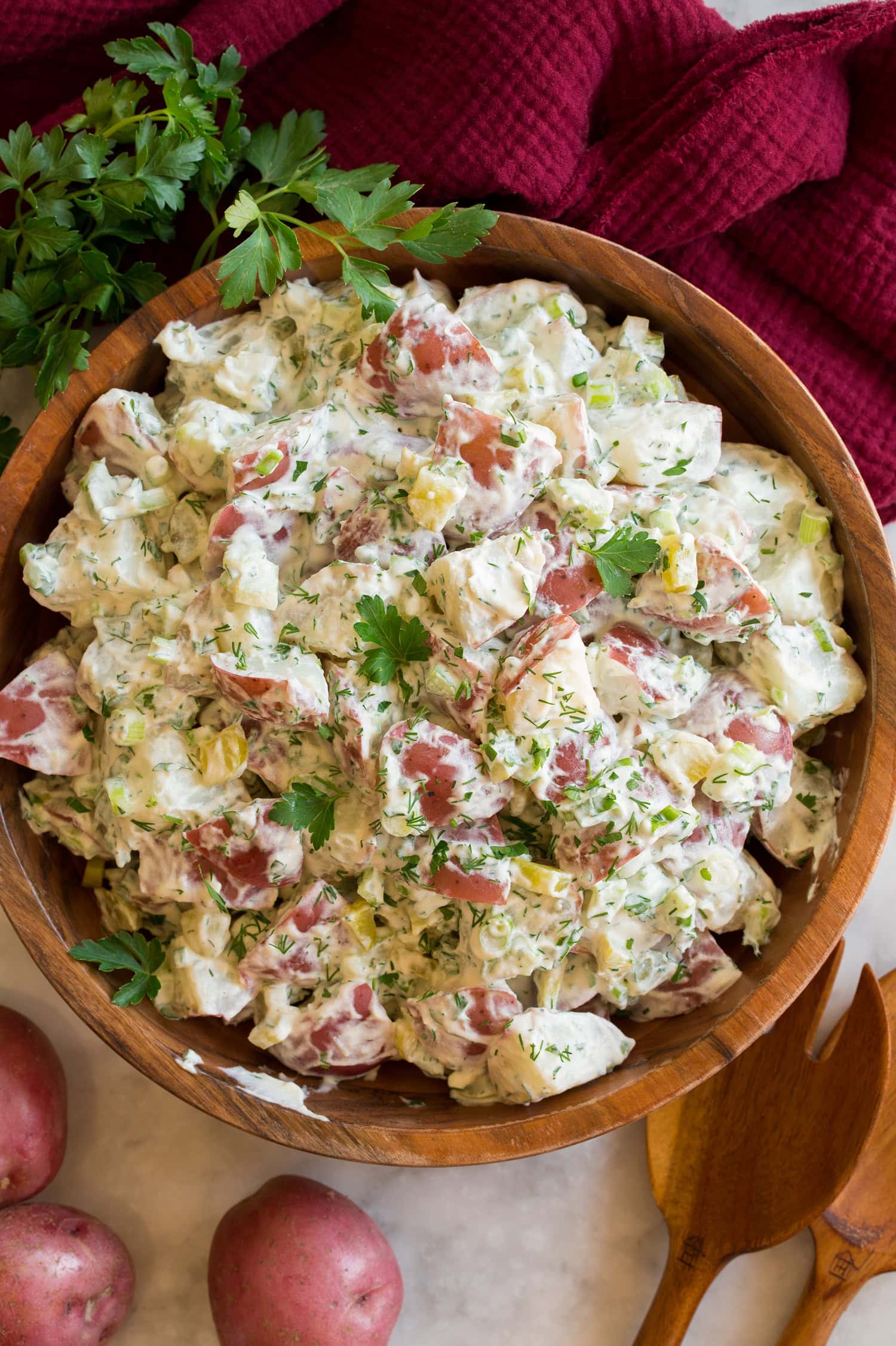 Overhead photo of red potato salad in a wooden bowl with a red cloth, whole potatoes, and parsley to the side. 