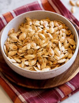 Roasted pumpkin seeds in a bowl set over a red and orange checkered cloth on a marble surface.