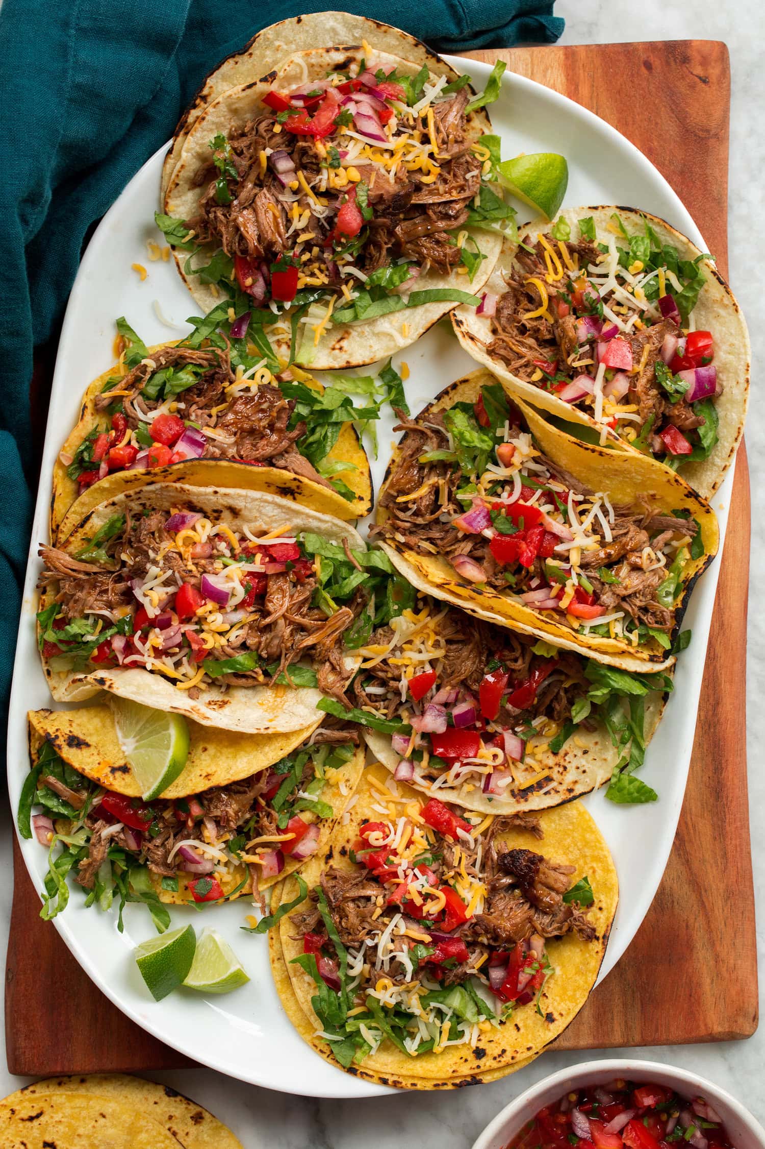 Overhead photo of eight shredded beef tacos on a white oval platter set on a wooden tray.