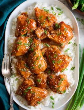 Slow cooker chicken thighs with sauce shown served over white rice on a white oval platter. Platter is on a marble surface with a blue cloth and green onions to the side.