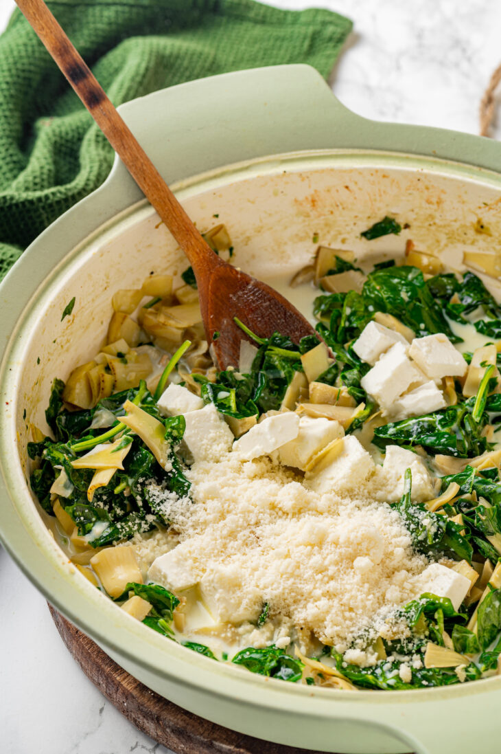 Grated cheese added to cream sauce with spinach and artichokes in a skillet, with a wooden spoon.